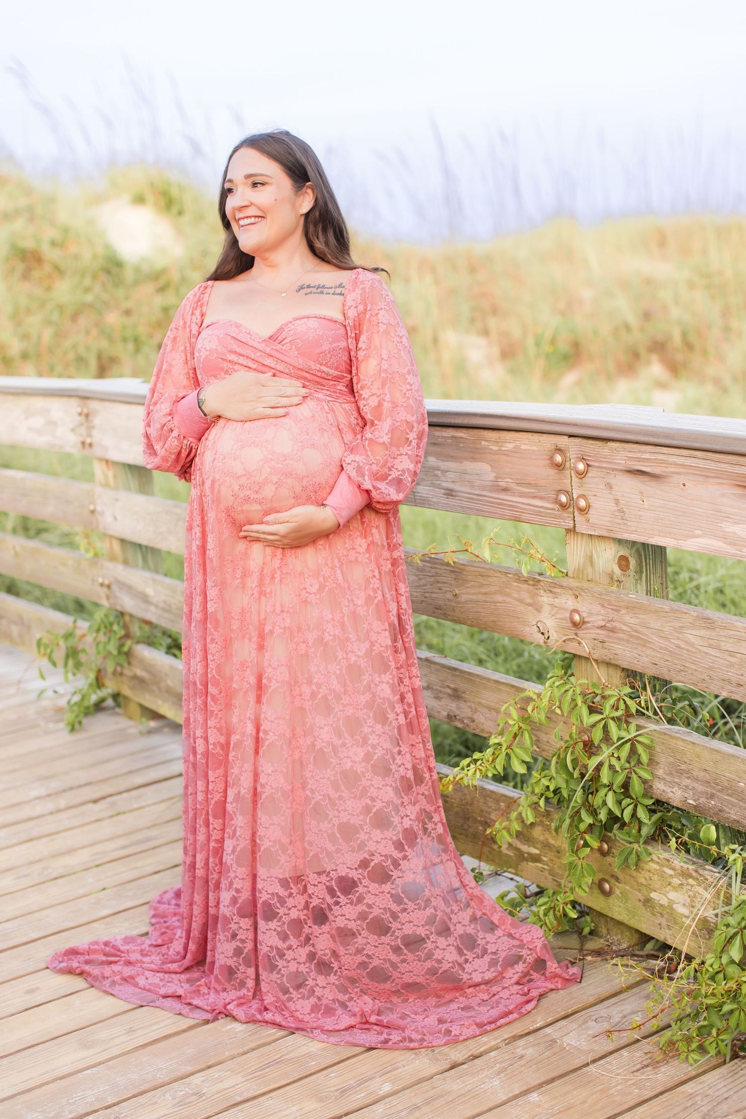 Smiling pregnant woman in elegant pink lace gown posing on a coastal wooden boardwalk.