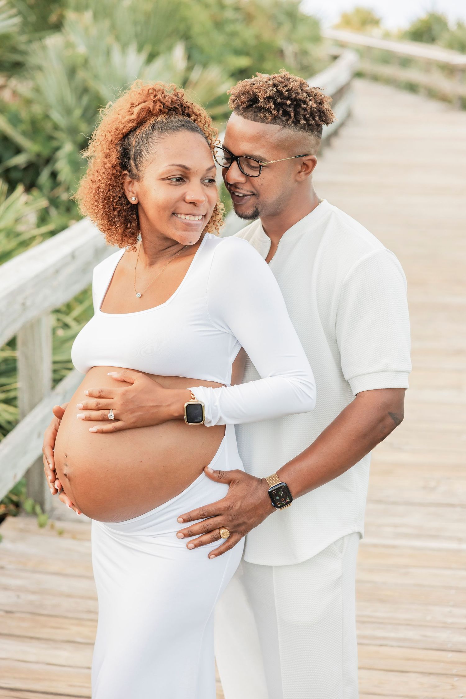 Expecting couple in matching white outfits on a boardwalk, partner lovingly holding pregnant womans baby bump.