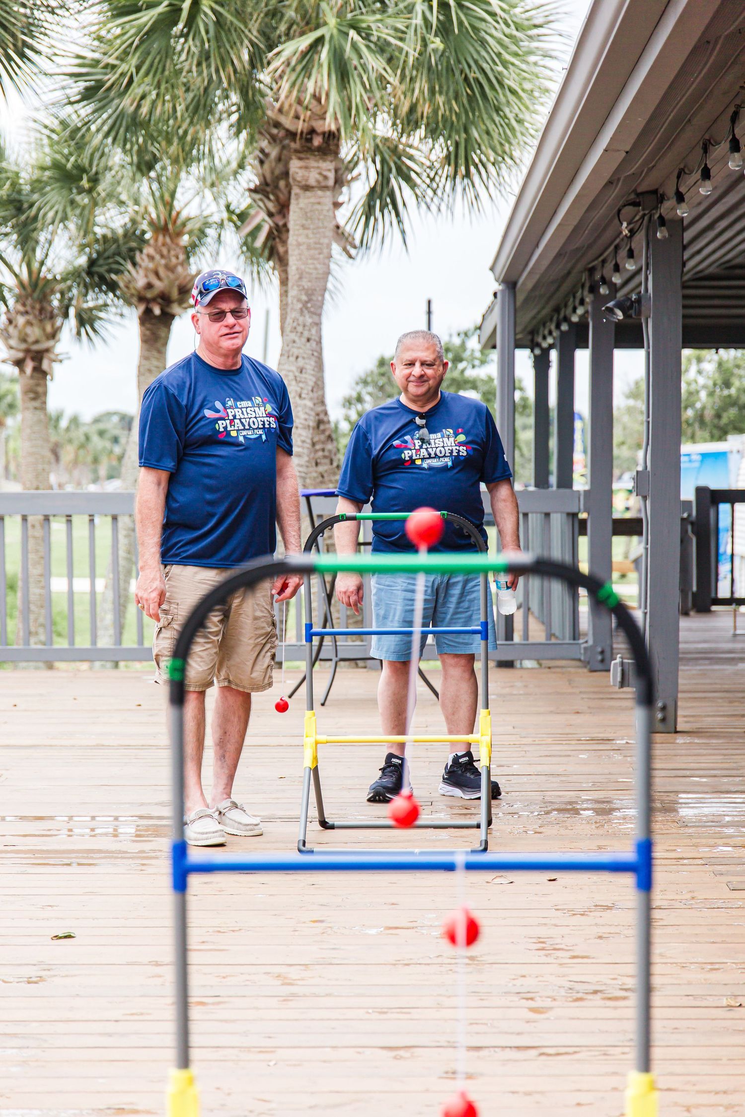 Two men in Prism Playoffs t-shirts play ladder toss on an outdoor wooden deck near palm trees.