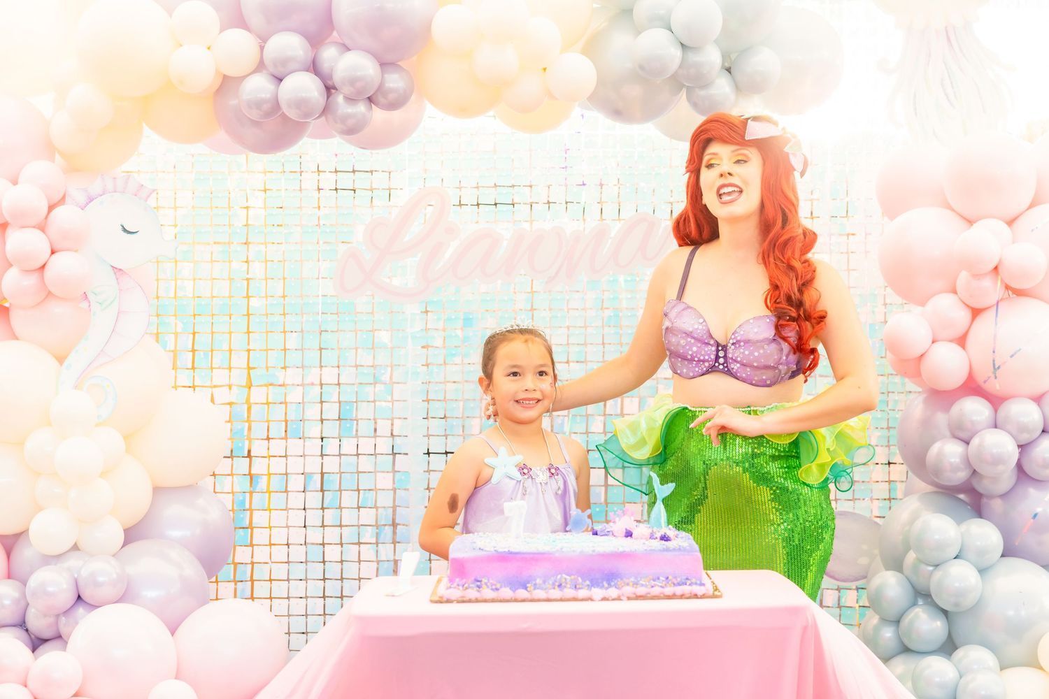 Birthday girl with Ariel mermaid character cutting purple mermaid cake at decorated party table.