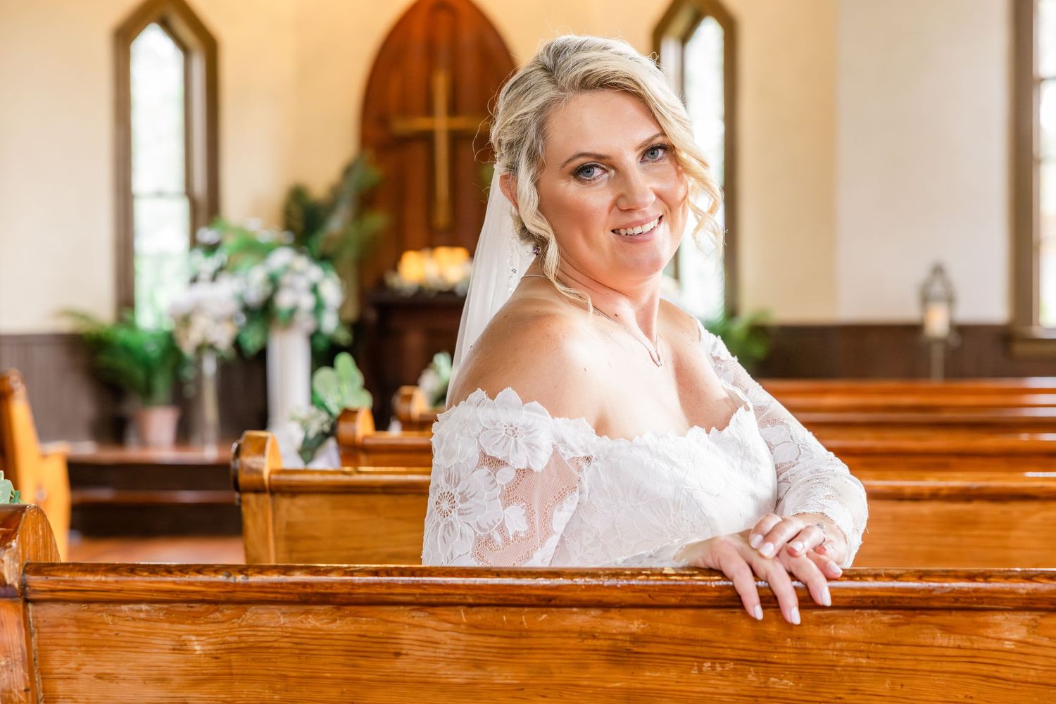 Smiling bride in off-shoulder lace gown sits in wooden church pew during bridal portrait.