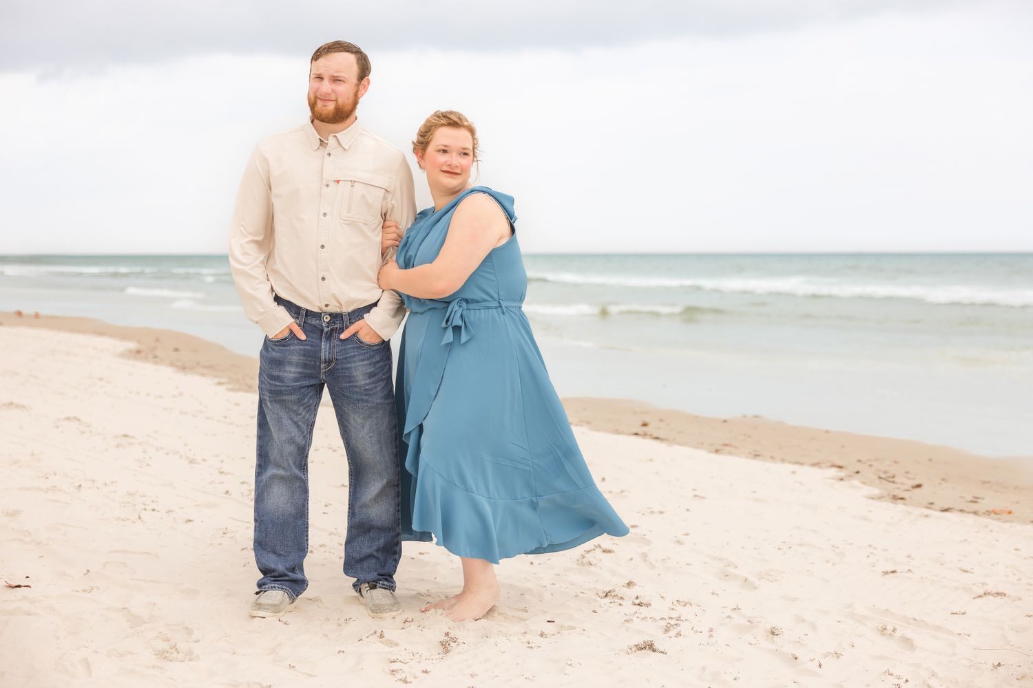 A couple holds hands and poses on a sandy beach with turquoise ocean waves behind them.