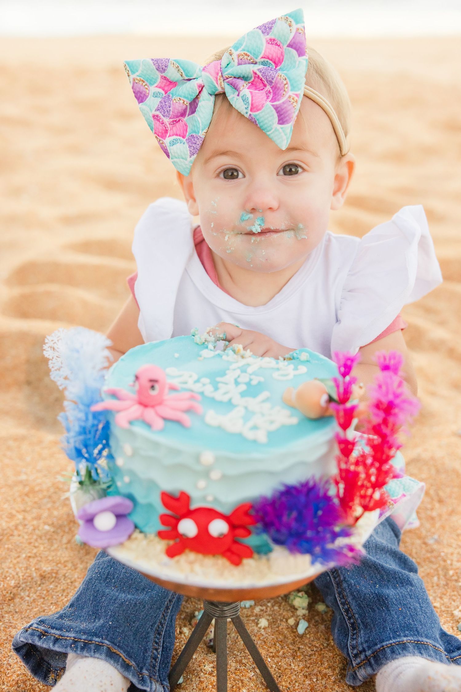 Baby girl in white top sitting with mermaid themed first birthday cake on beach.