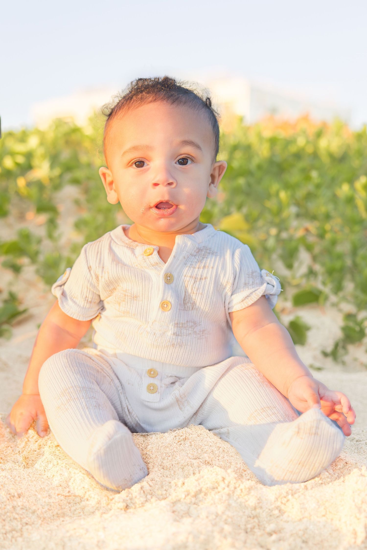 Adorable baby in white linen outfit sitting on sandy beach in warm sunlight.