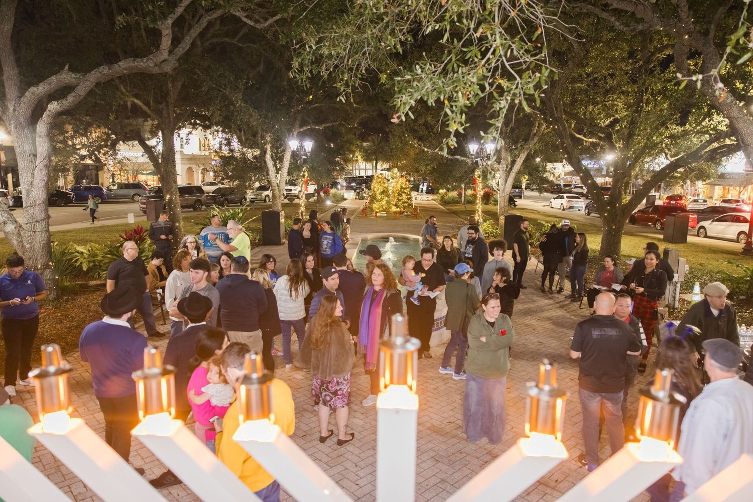 Crowd gathers outdoors at night around illuminated Hanukkah menorah under oak trees.