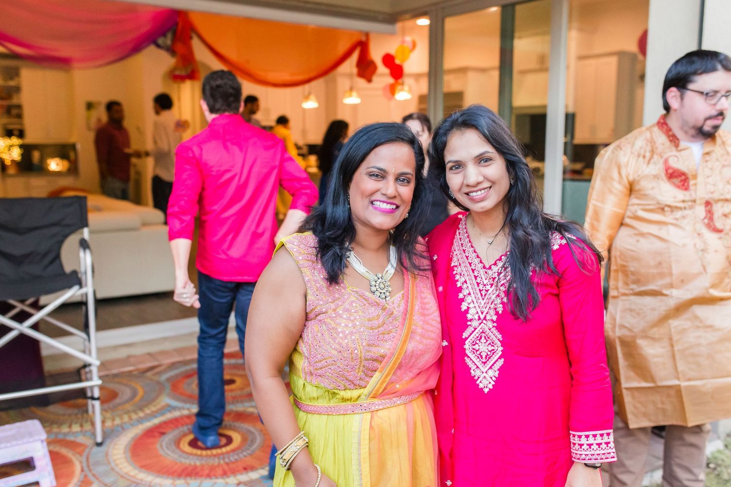 Two women in vibrant pink and yellow Indian attire smile together at a festive celebration.