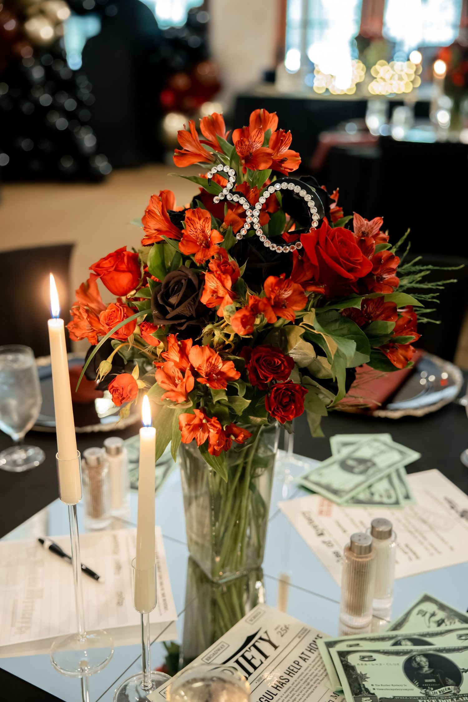 Lush orange and red floral centerpiece with pearl 30 number candles and money props on dark table.