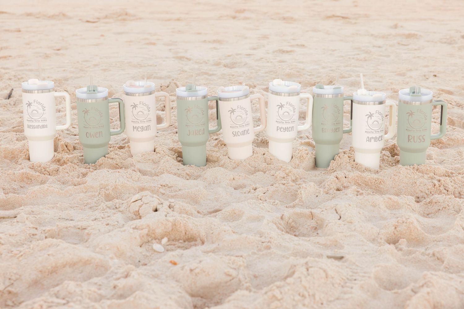 Six personalized tumblers with lids lined up in the sand on a sunny beach.