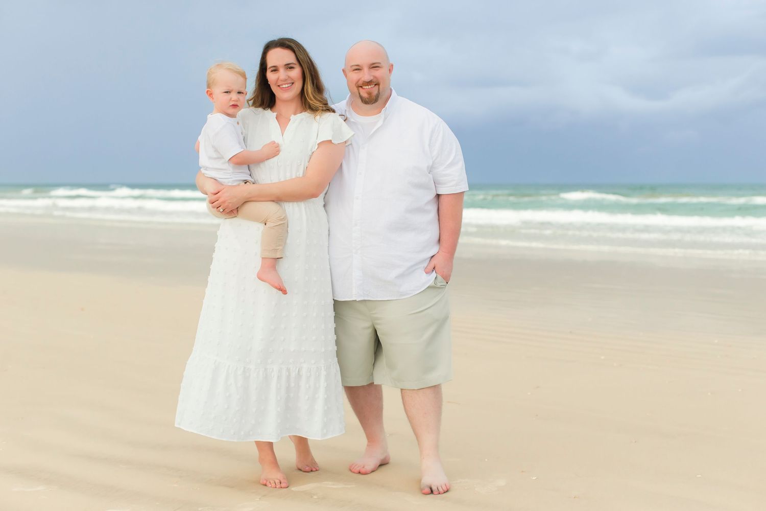 Family of three posing on a sandy beach, mother in white dress holding baby with bald father beside her.