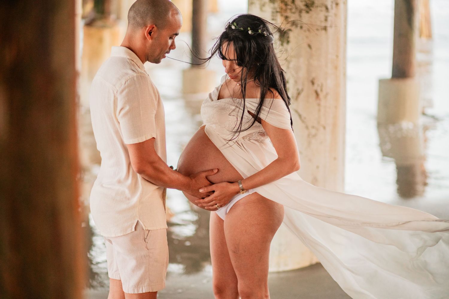Expectant couple sharing intimate moment under pier, man gazing at pregnant partners belly.