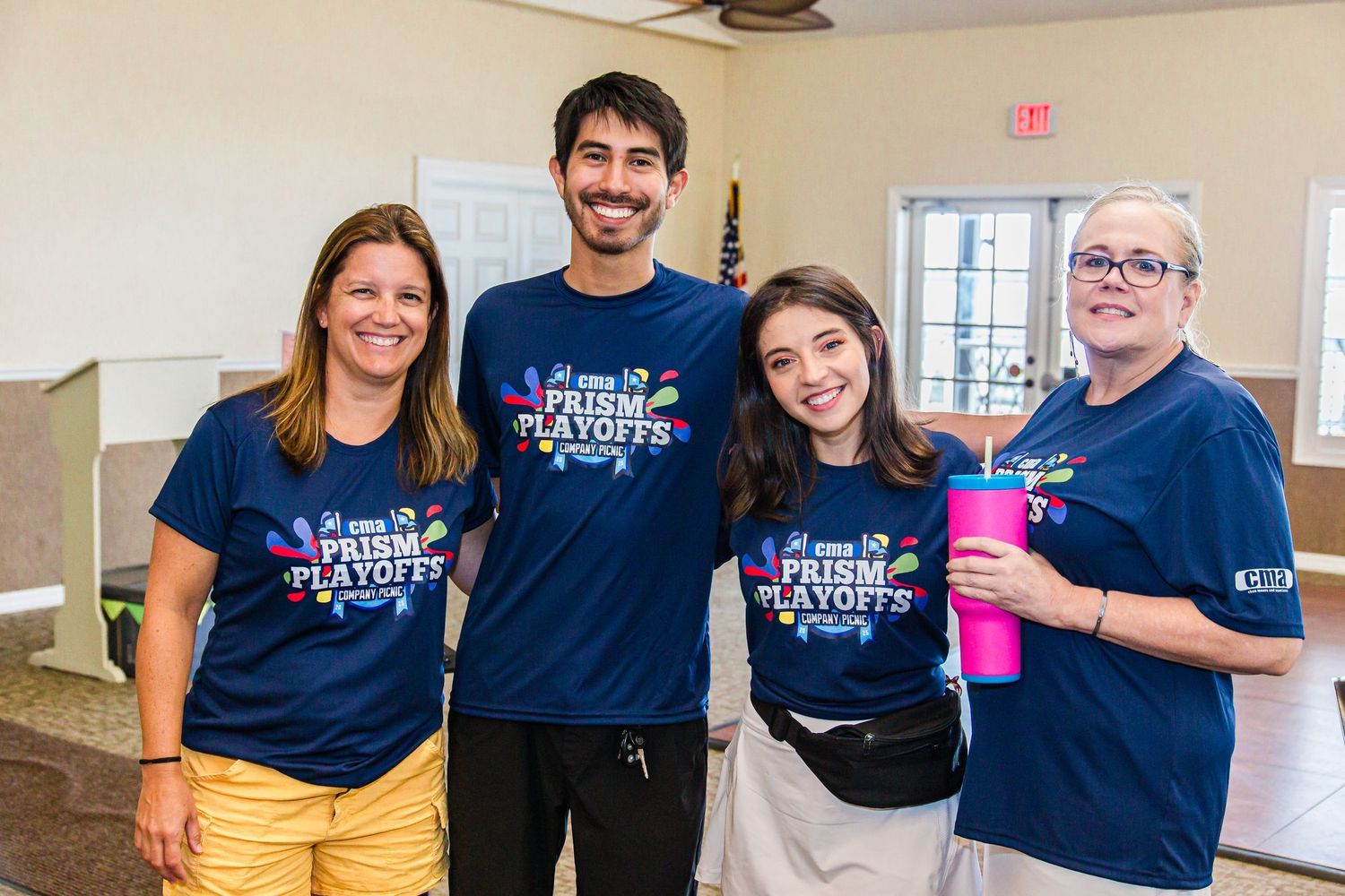Four smiling volunteers wearing Prism Playoffs t-shirts pose together indoors at a community event.