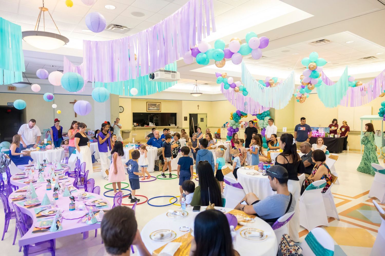 Children and families celebrate at a colorful Jasmine themed birthday party with purple and teal balloon decorations.