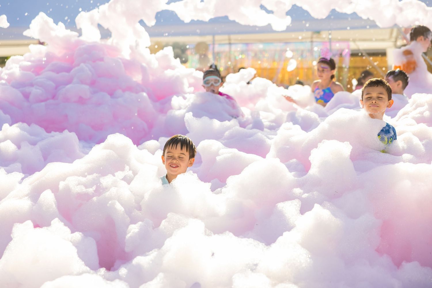 Children laughing and playing in colorful pink and white foam bubbles at outdoor party.
