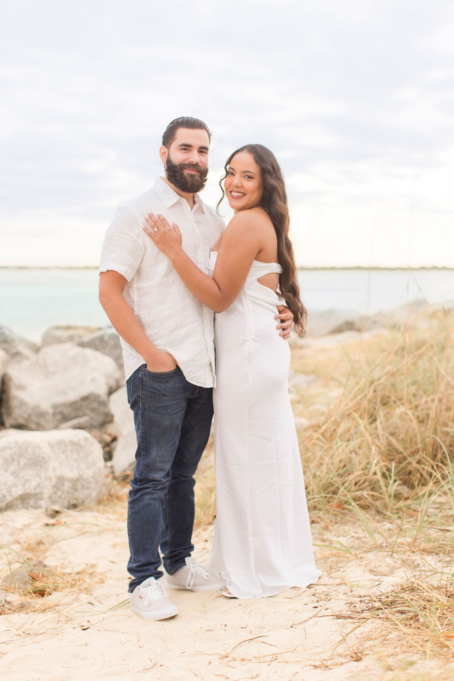 Couple embracing on sandy beach path with rocks and ocean behind them.