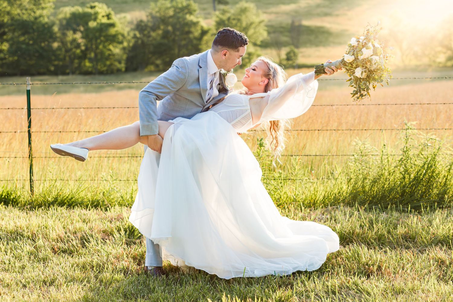 Groom lifting bride in white ball gown during golden hour in lush green farm field.