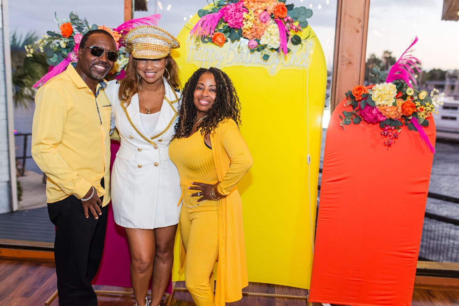 Three friends pose together in front of a vibrant yellow lemon backdrop at an outdoor celebration.