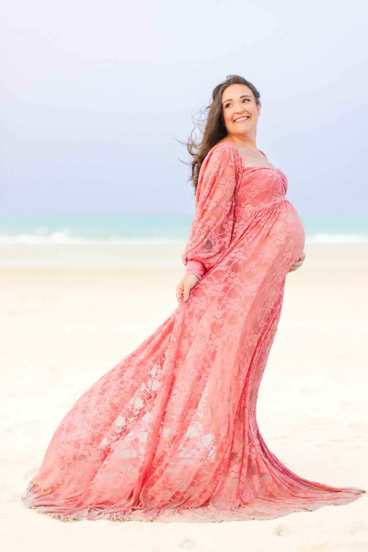 Pregnant woman in flowing pink lace gown on sandy beach, cradling baby bump, with ocean backdrop.
