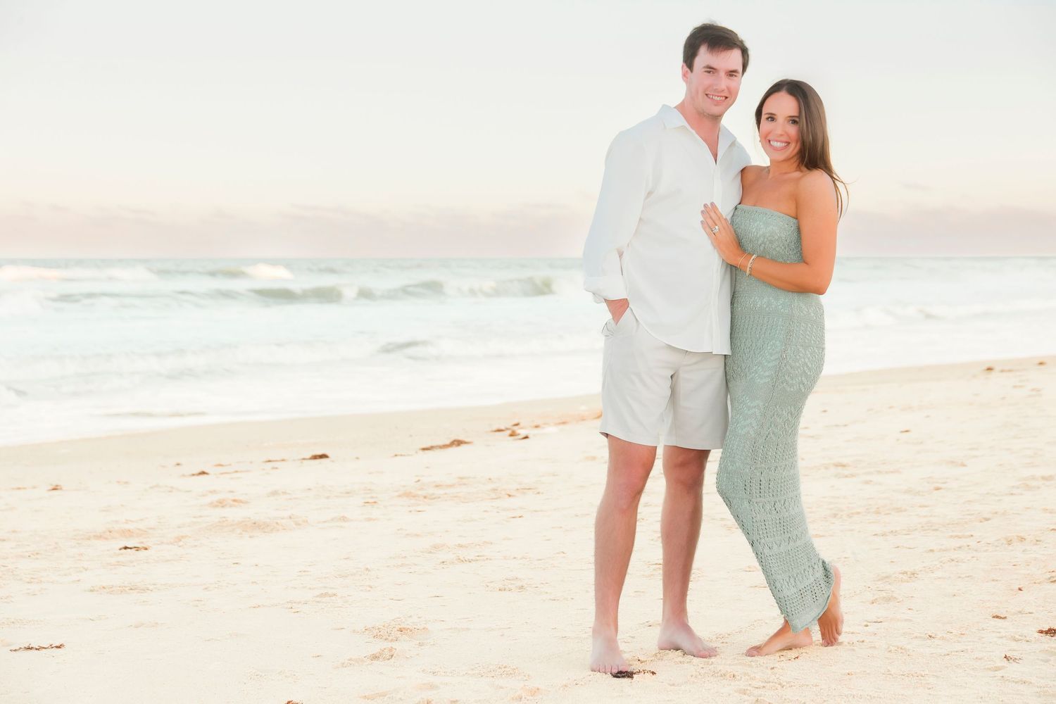 Young couple posing together on a sandy beach at sunset, man in white shirt and woman in green jumpsuit.