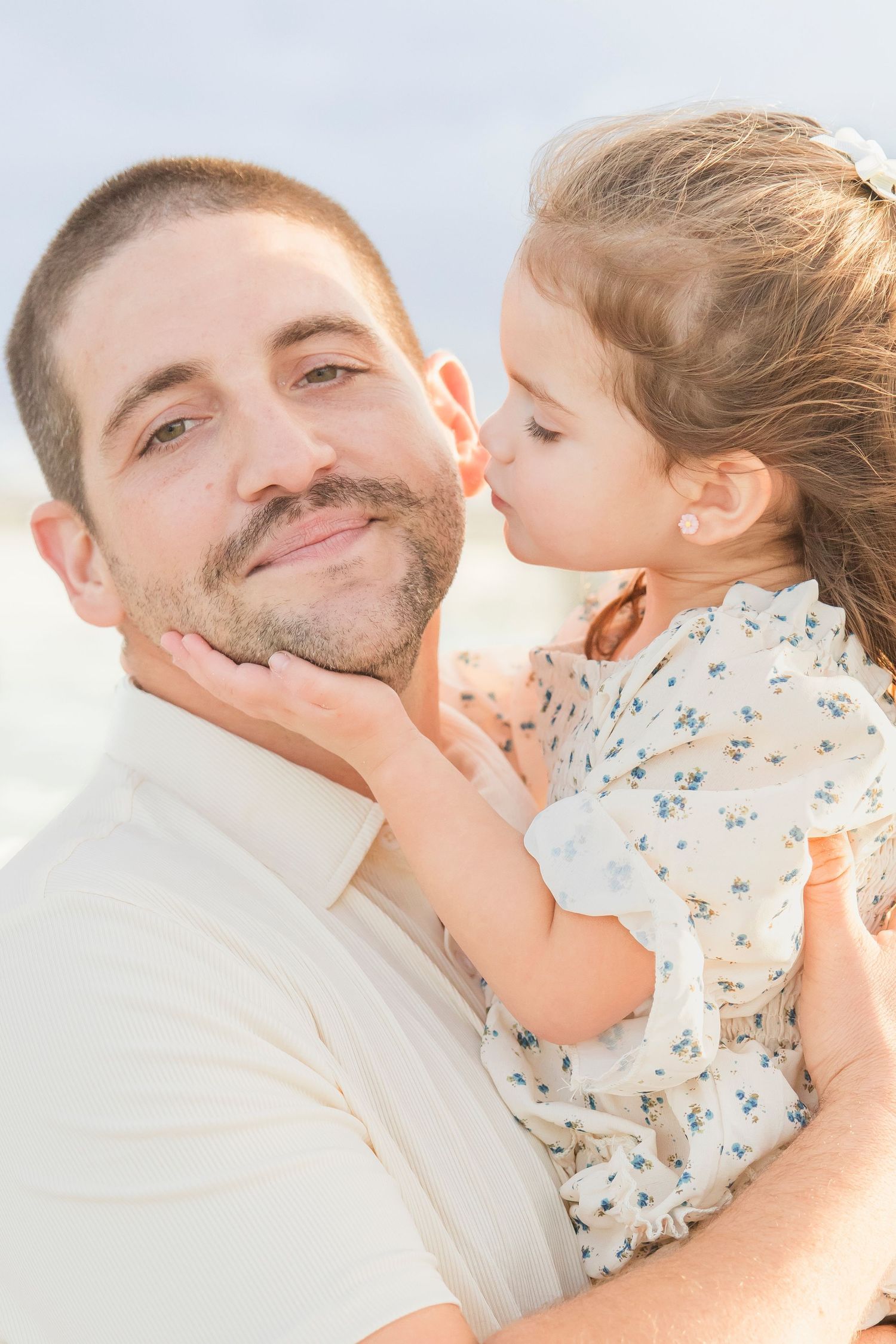 A young girl kisses a smiling man on the cheek while being held at a beach.
