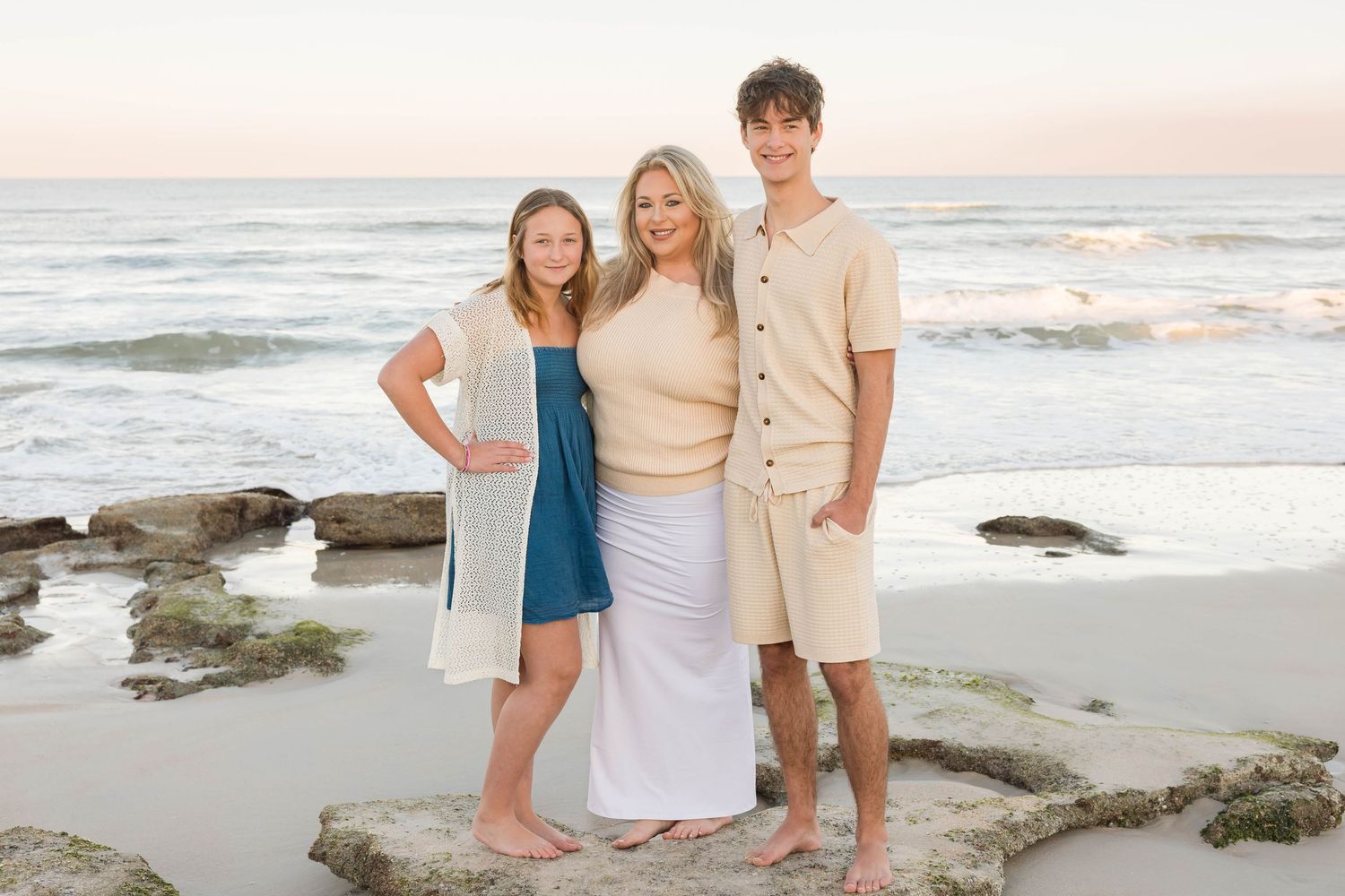 Mother posing with teenage son and daughter on rocky beach at sunset, all smiling.
