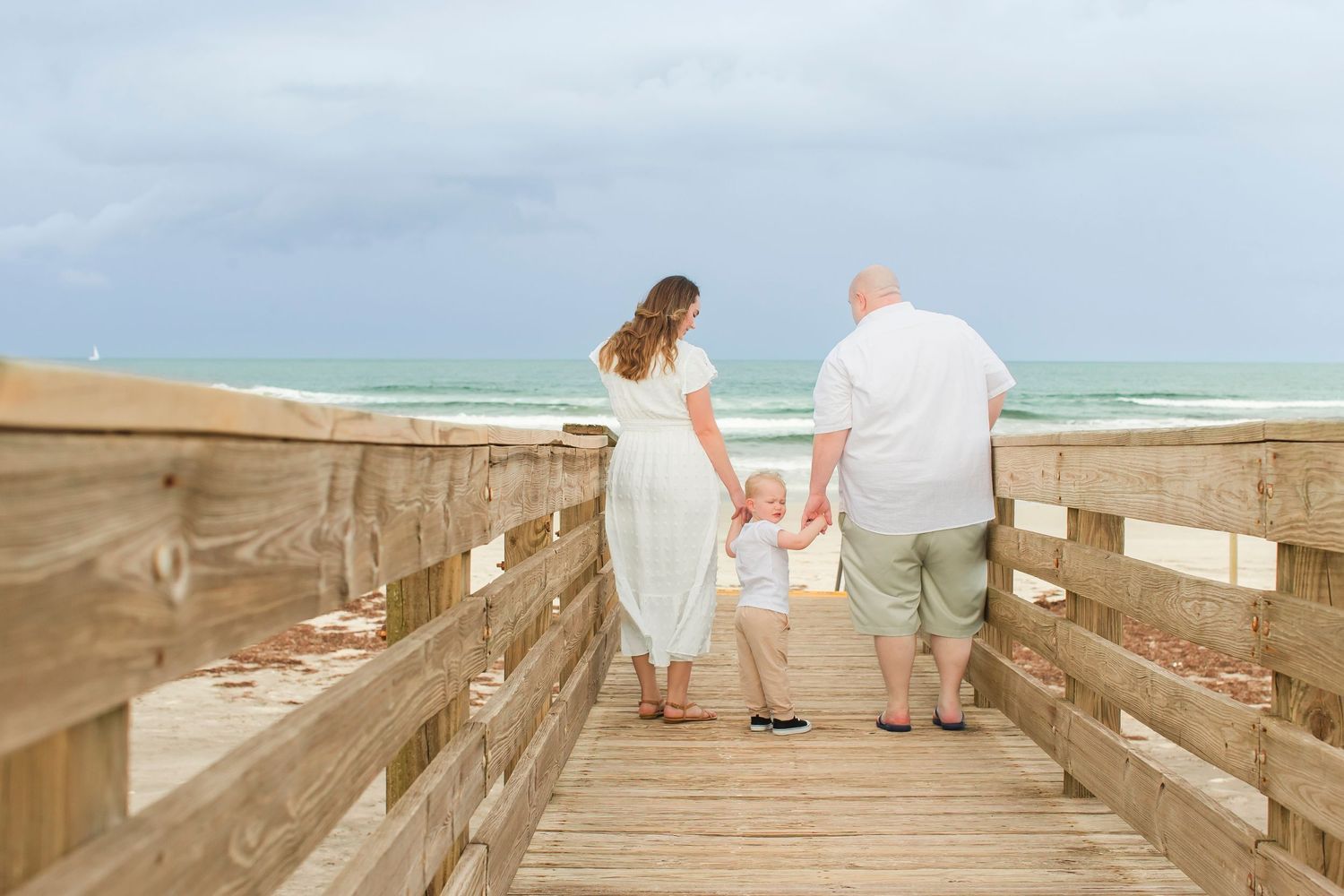 Family of three walking on a beach boardwalk toward the ocean, parents holding toddler's hands.