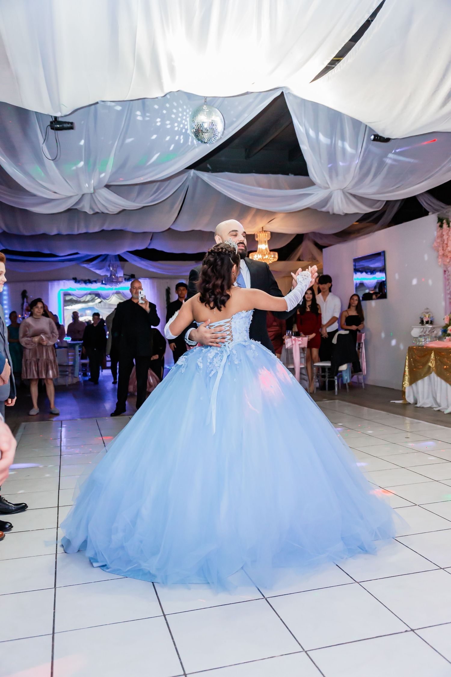 A young woman in a light blue ball gown dances with a partner at a quinceañera celebration.