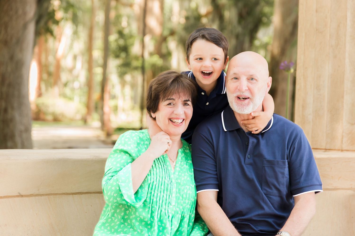 Happy grandparents sitting outdoors with their young grandson smiling between them in a sunny park setting.