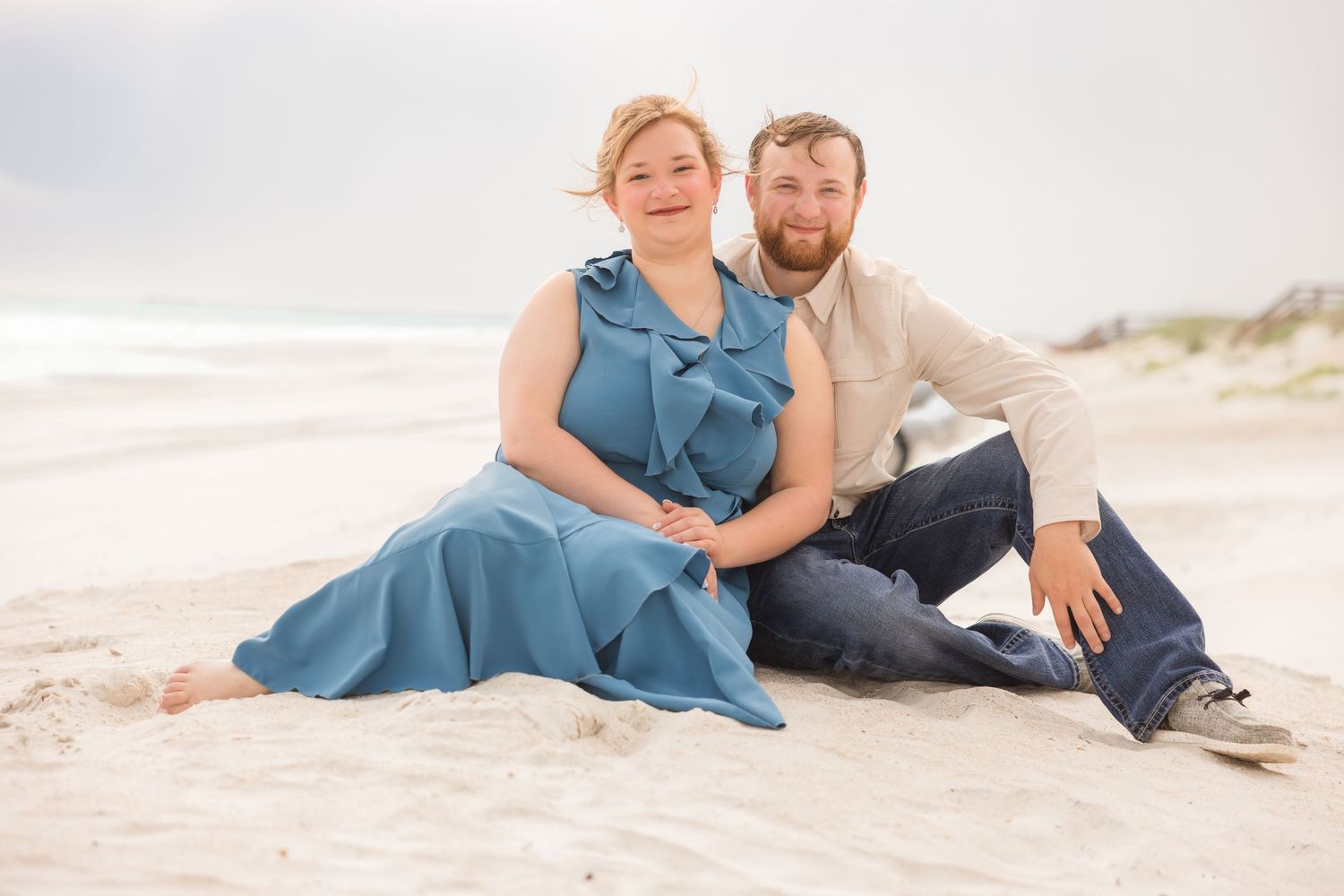 A smiling couple sits together on a sandy beach, woman in blue dress leaning on man.
