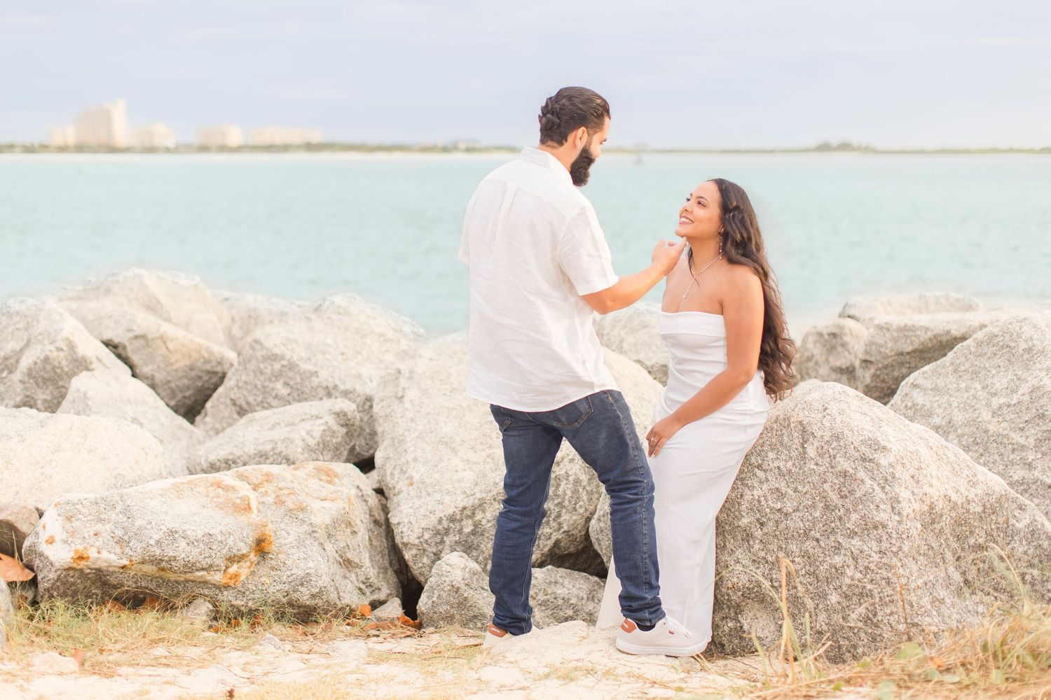 Couple holding hands near large rocks with turquoise ocean in background.
