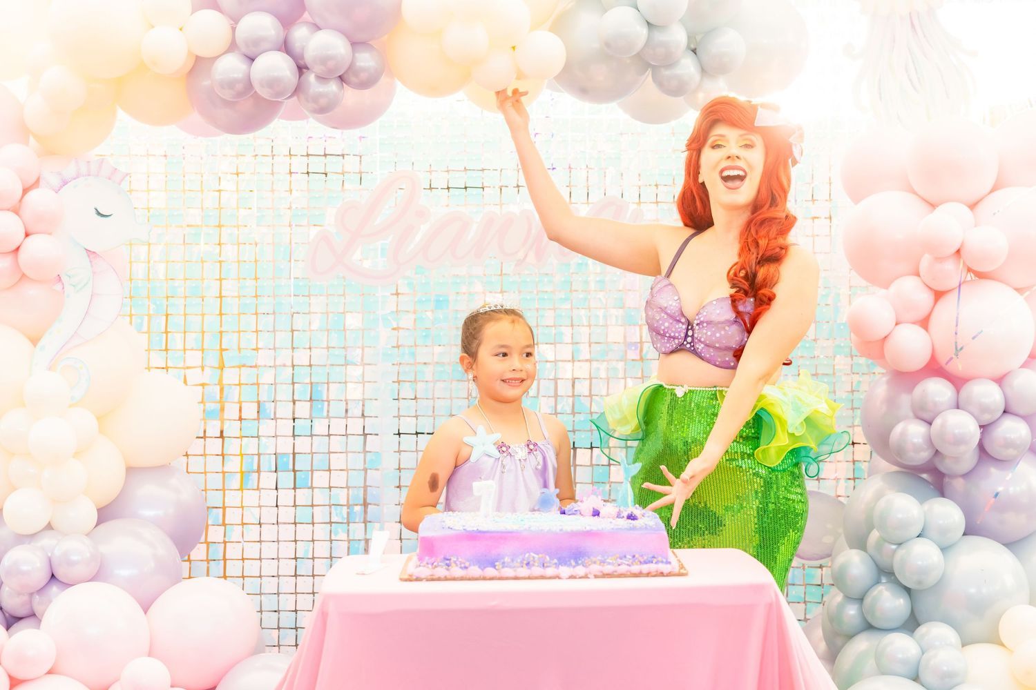 A little girl celebrates her birthday with an Ariel Little Mermaid princess character beside a cake surrounded by pastel balloons.
