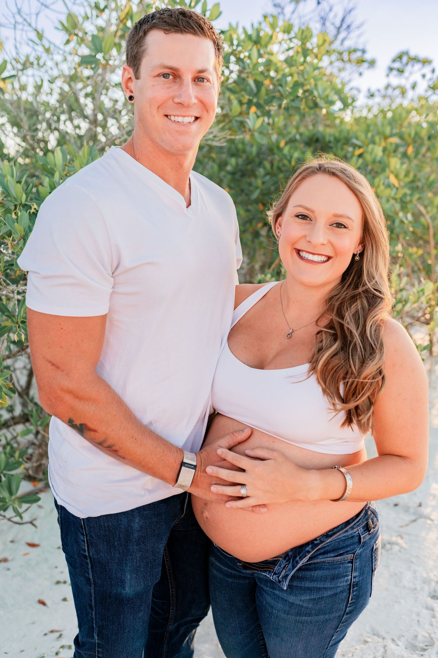 Happy couple smiling at camera on sandy beach, man embracing pregnant woman from behind.