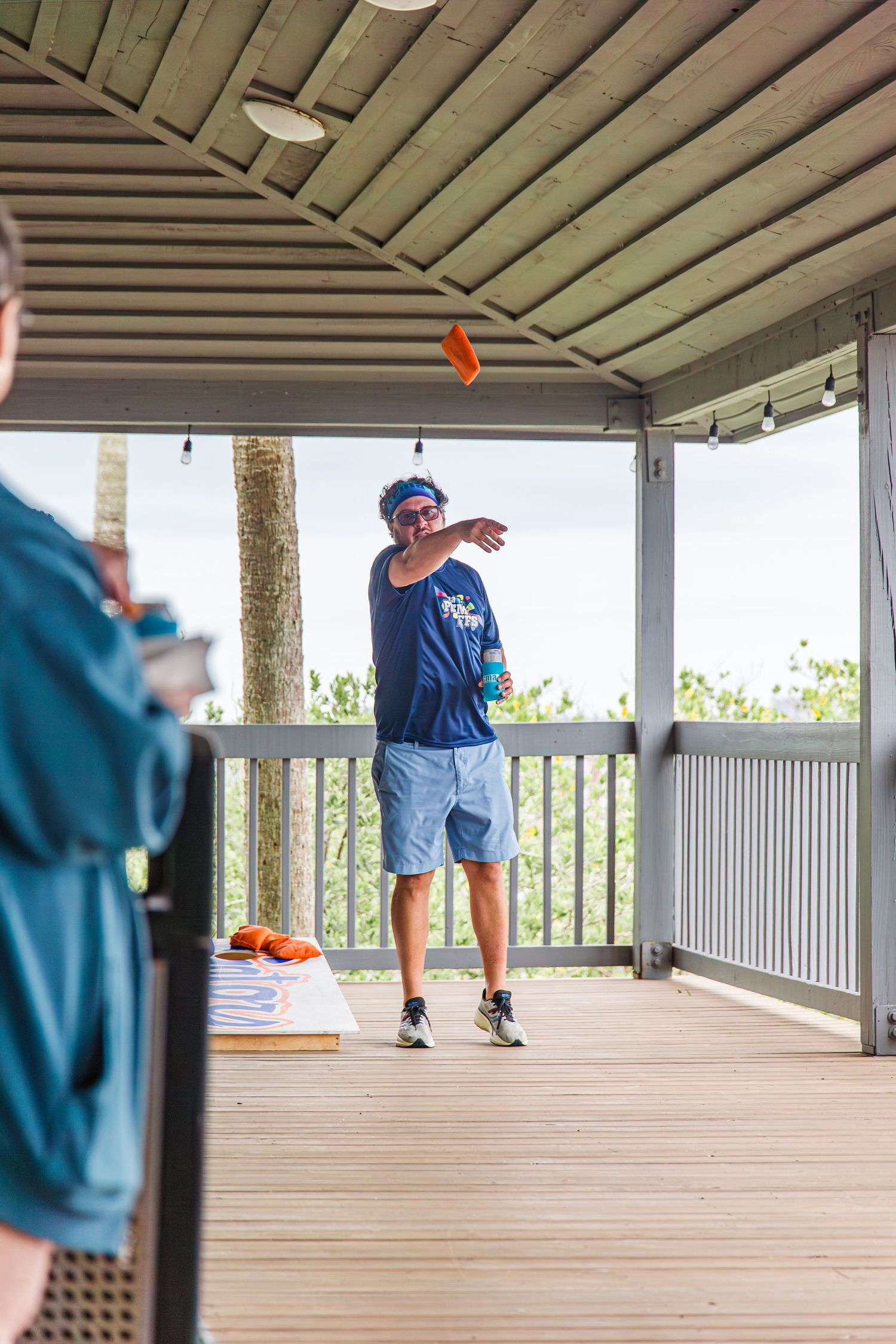 Man tossing a cornhole bag on a covered wooden deck during an outdoor game event.