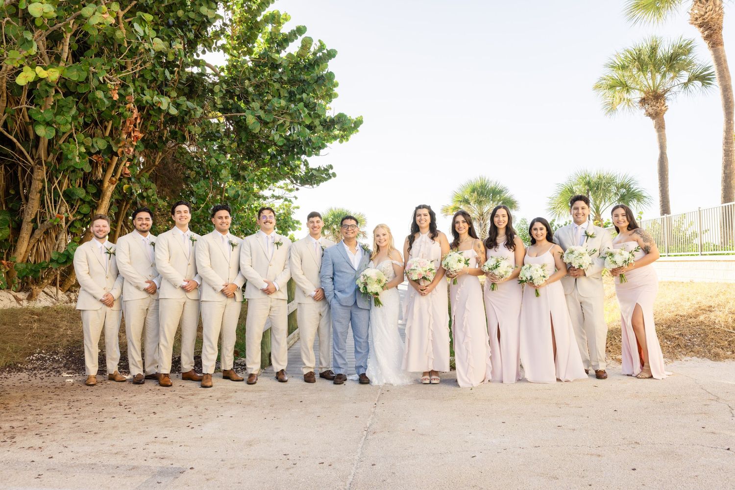 Wedding party poses outdoors in neutral tones with groomsmen in tan suits and bridesmaids in blush dresses.