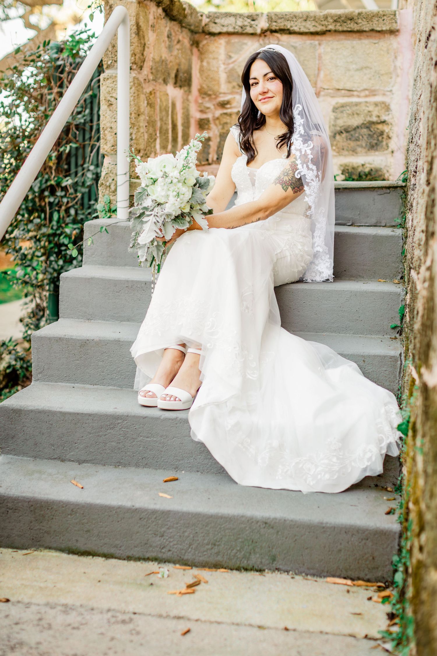 Brunette bride in lace gown and veil sitting on stone steps holding white floral bouquet.