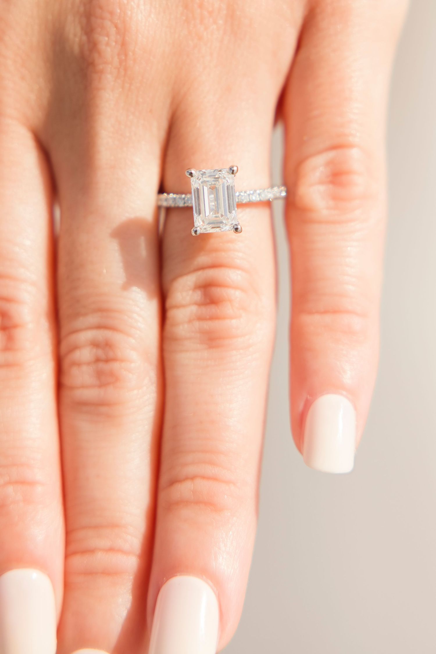 Elegant emerald cut diamond solitaire engagement ring with pavé band on a woman's hand with nude nails.