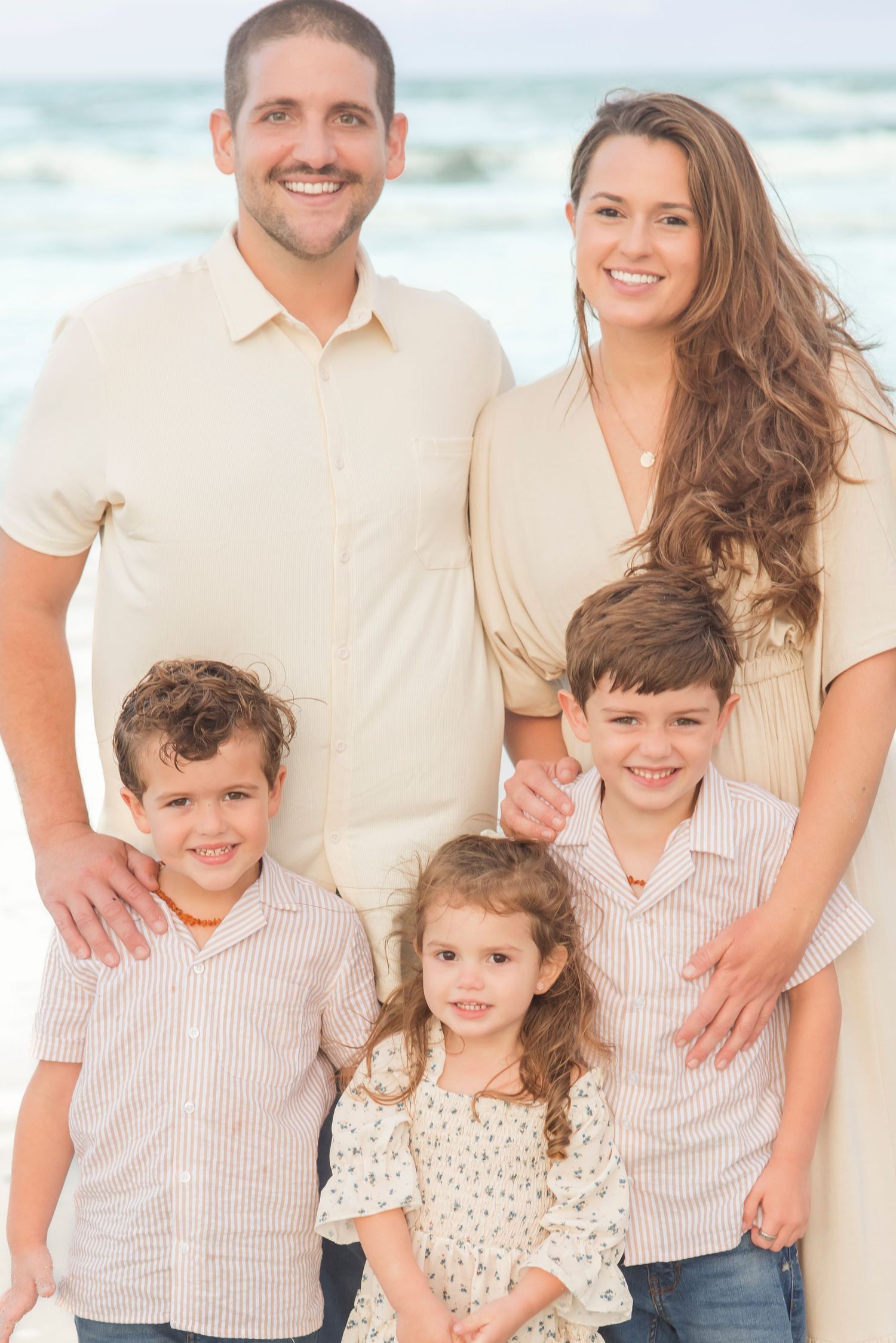 Happy family of five posing together on a sunny beach, parents smiling with three young children in coordinating outfits.