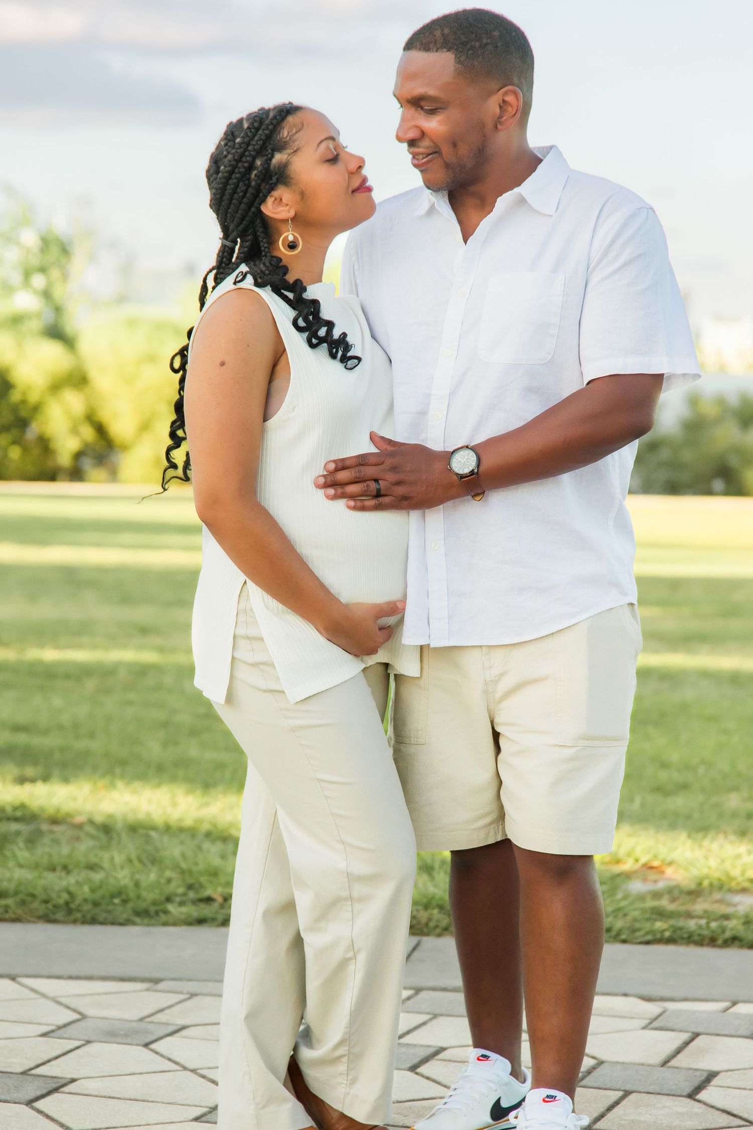 Expecting couple in matching white outfits sharing a tender moment outdoors, man gently touching pregnant woman's belly.
