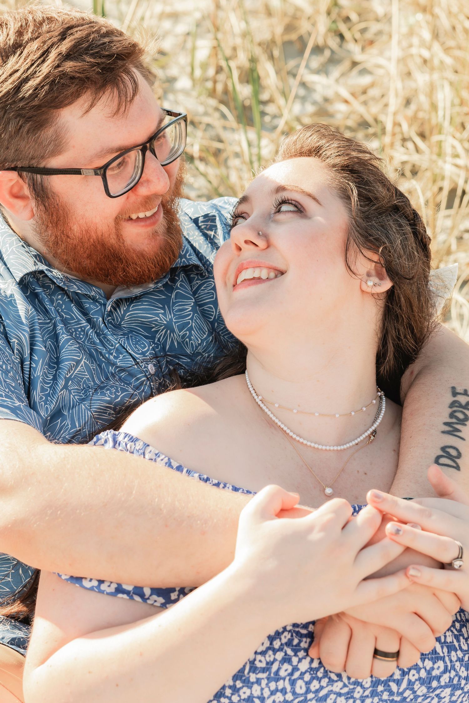 Happy couple lying in grass holding hands, woman looking up at bearded man wearing glasses.