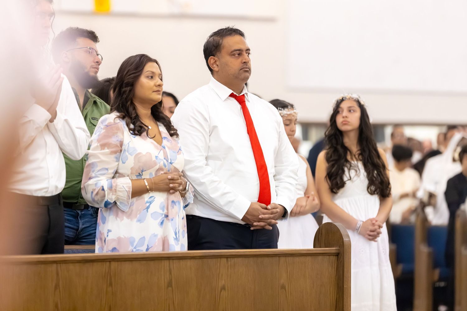 A man in a white shirt and red tie stands with a woman in a floral dress during a church ceremony.