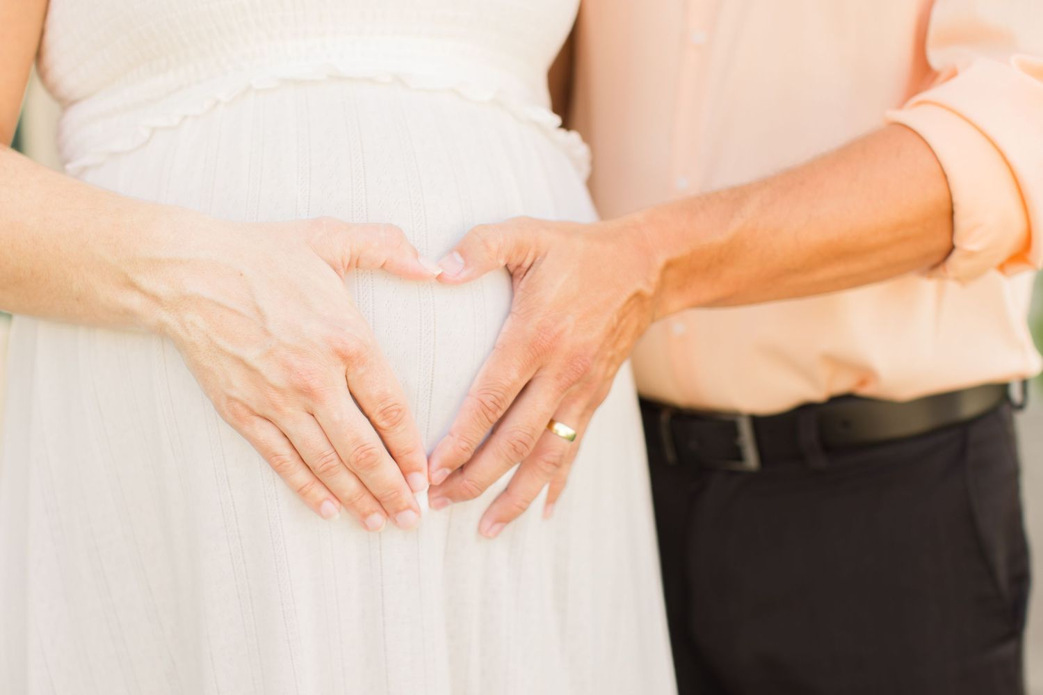 Couple forming heart shape with hands on pregnant belly, maternity photo closeup detail shot.