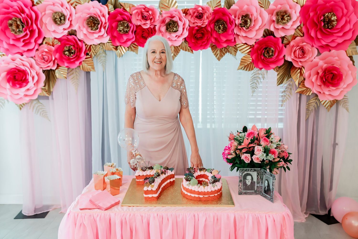 Woman standing beside a number 70 birthday cake under a pink paper flower arch decoration.