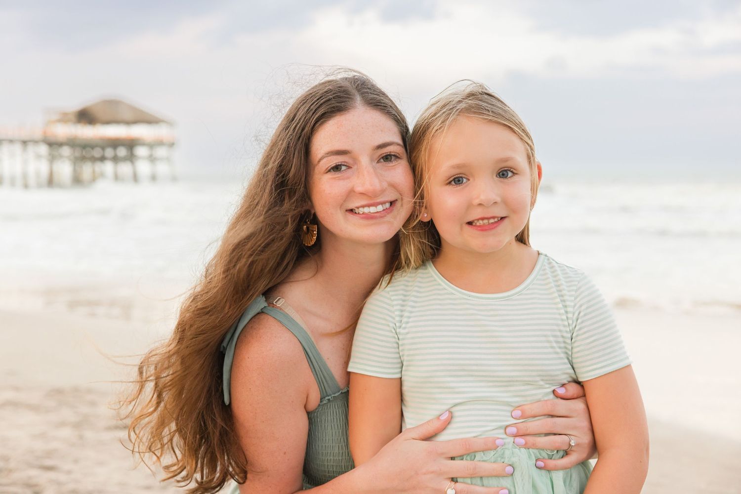 Teen girl with long brown hair hugging young blonde girl smiling on a sunny beach.