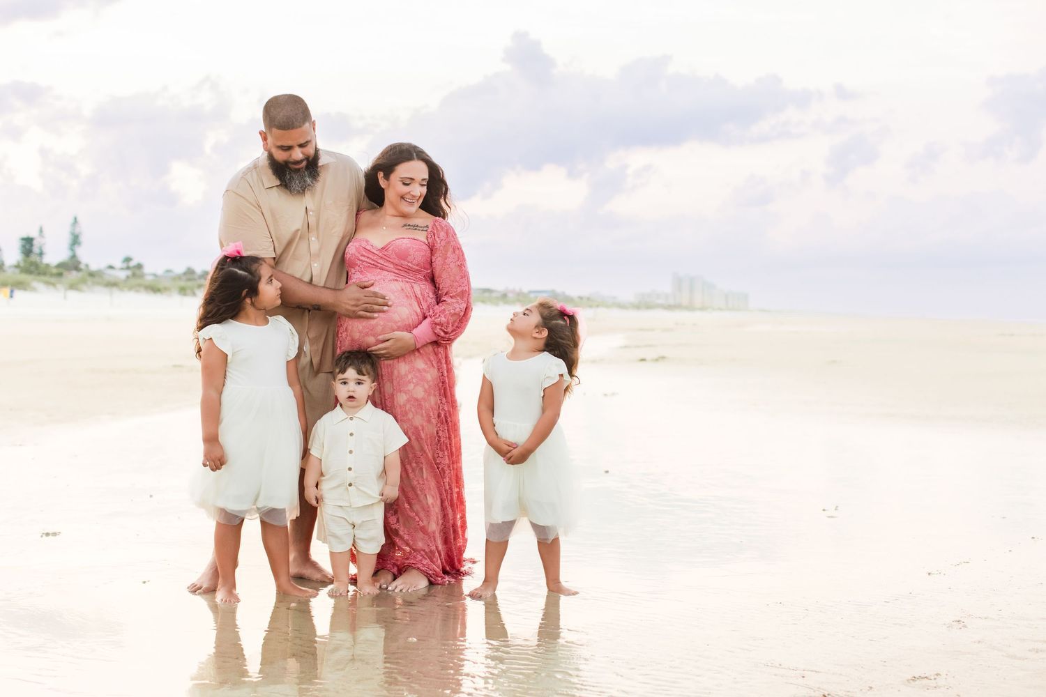Pregnant woman in pink gown with family on beach, husband and three children surrounding her bump.