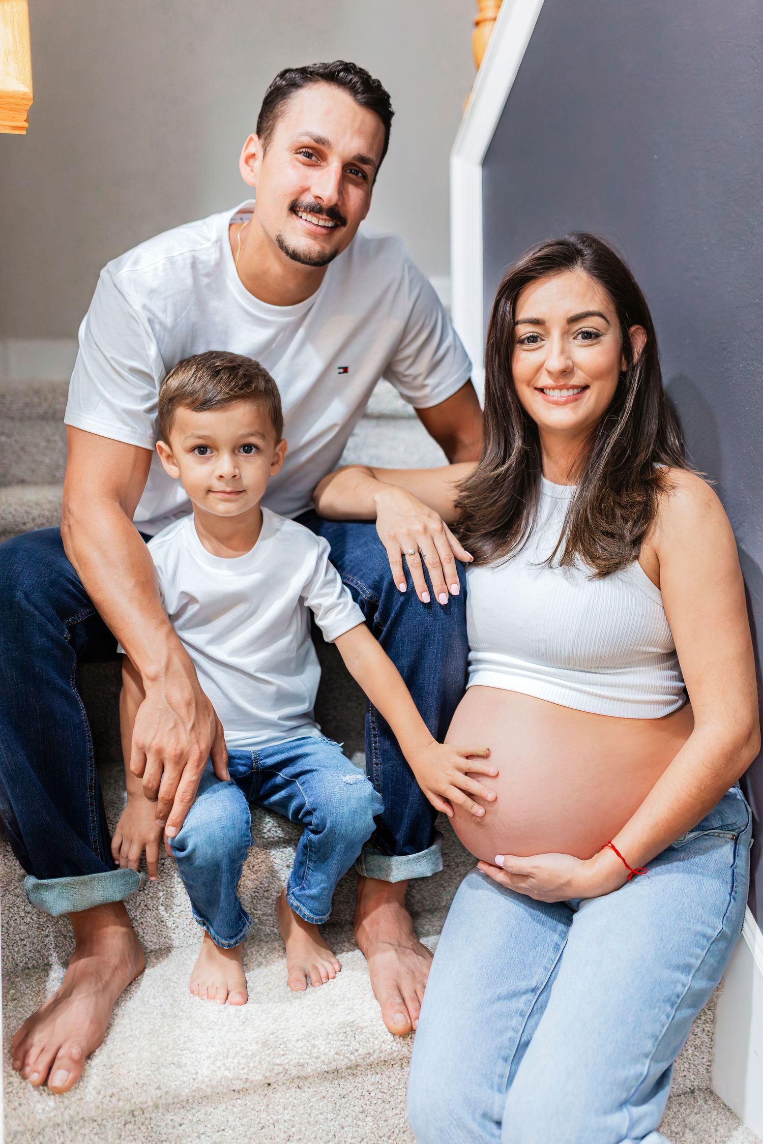 Smiling family with pregnant mother, father, and young son sitting together on home staircase.