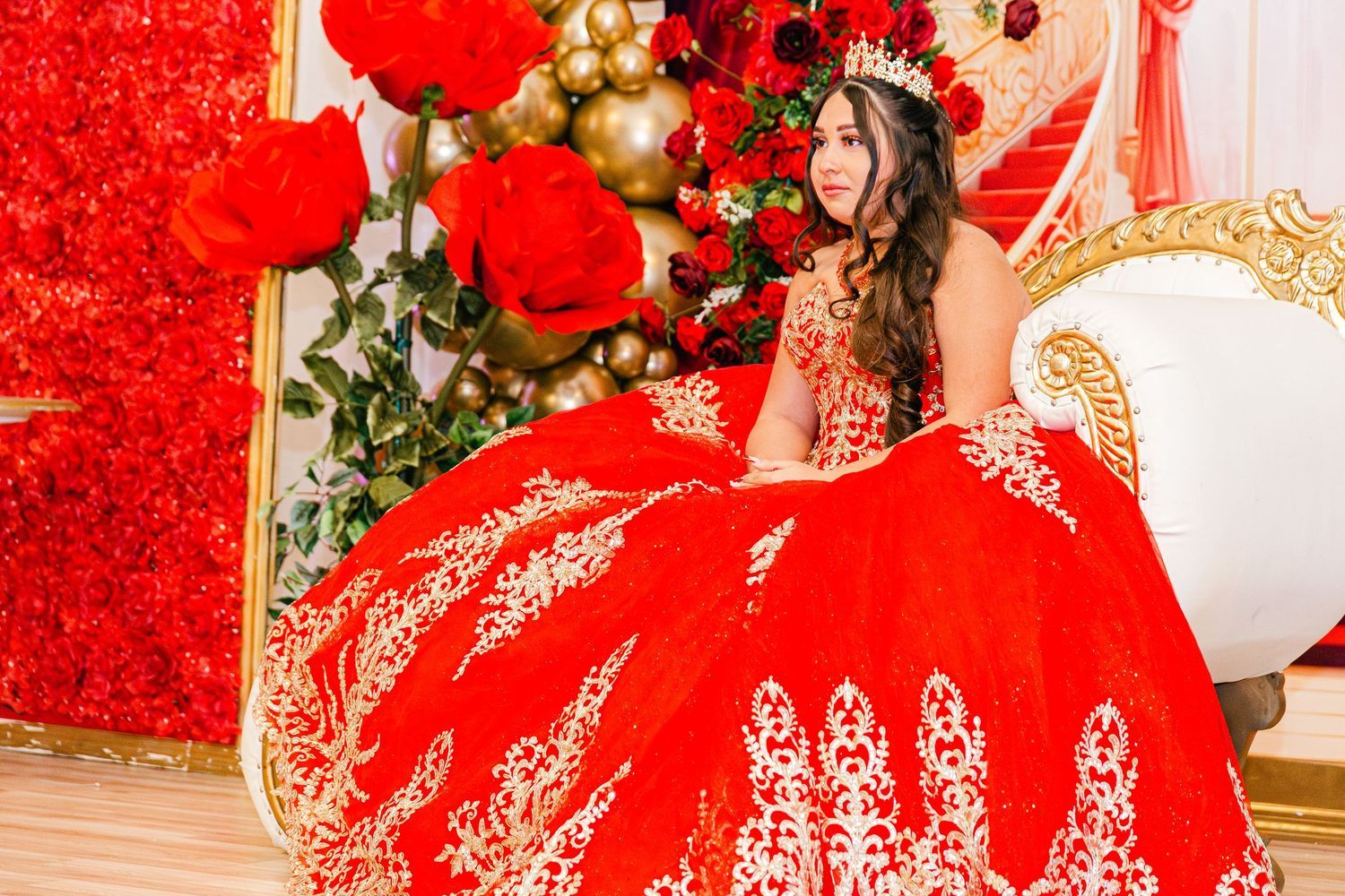 Young woman in stunning red and gold quinceañera ball gown posing with floral decorations.