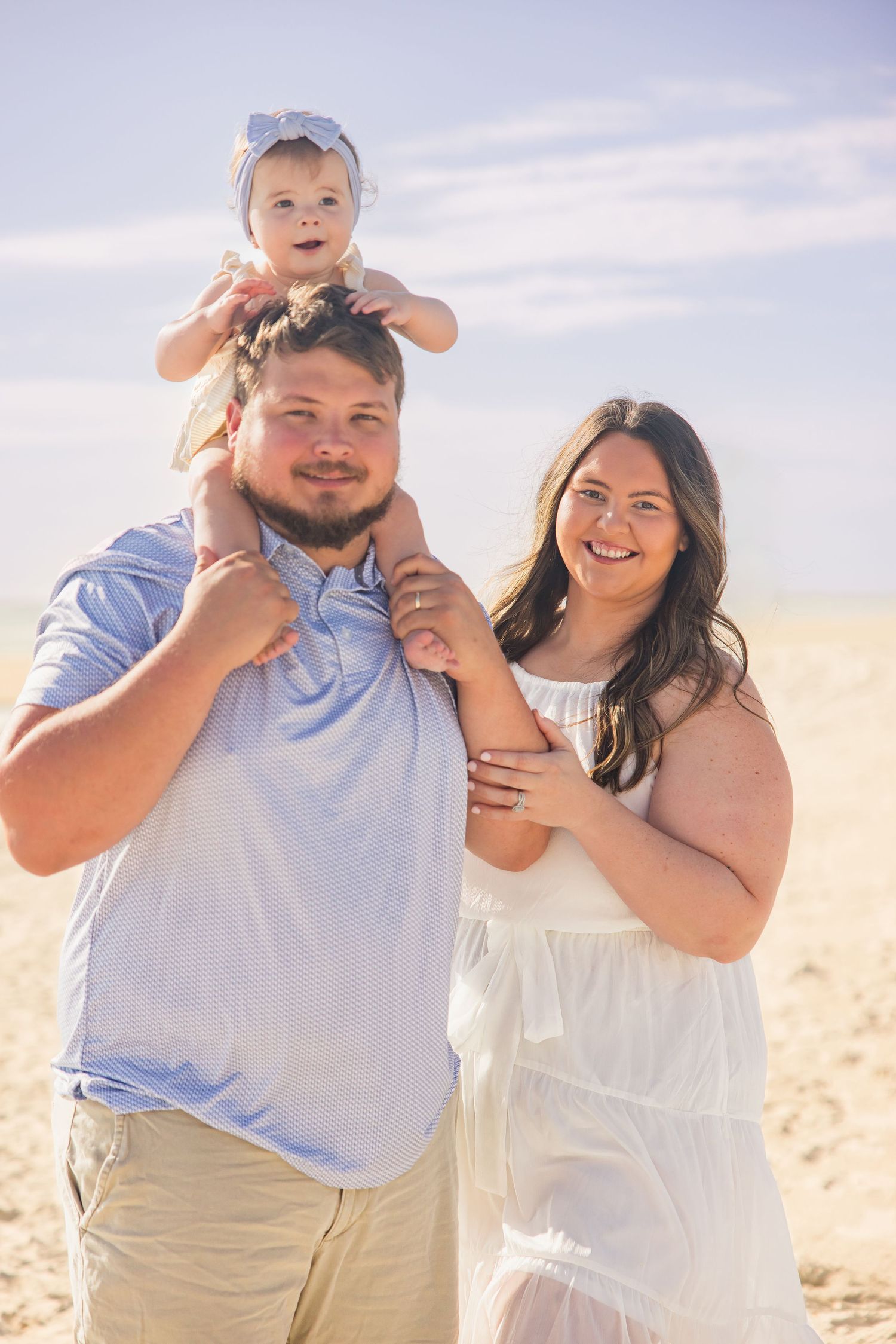 Happy family of three with baby on fathers shoulders smiling on sunny beach.