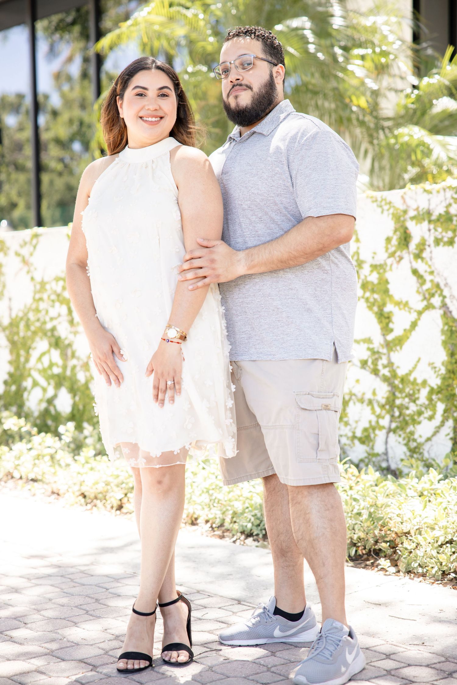 Couple posing outdoors, woman in white dress and man in gray polo shirt near tropical greenery.