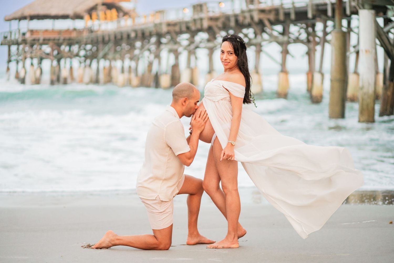 Man kneeling to kiss pregnant womans belly on beach near pier, romantic maternity shoot.