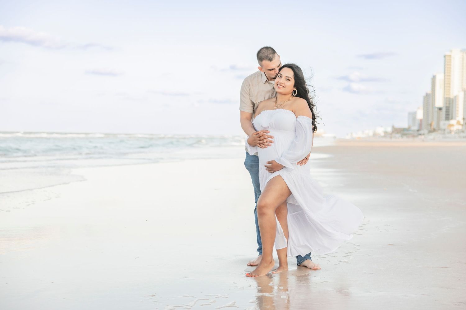 Expecting couple embracing on a beach at sunset, woman in flowing white maternity dress with city skyline behind.