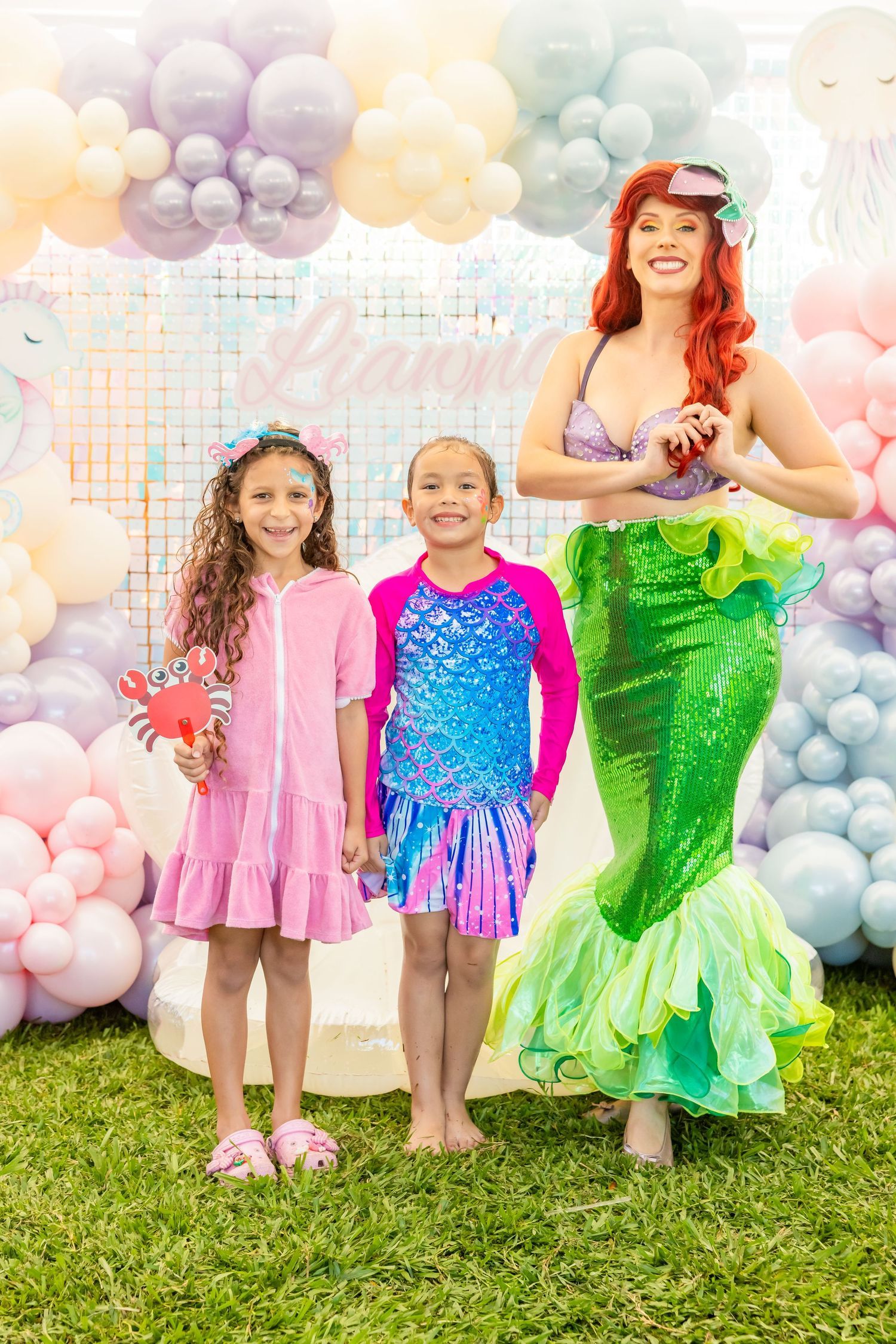 Two girls in swimsuits posing with Little Mermaid Ariel character at mermaid-themed birthday party.